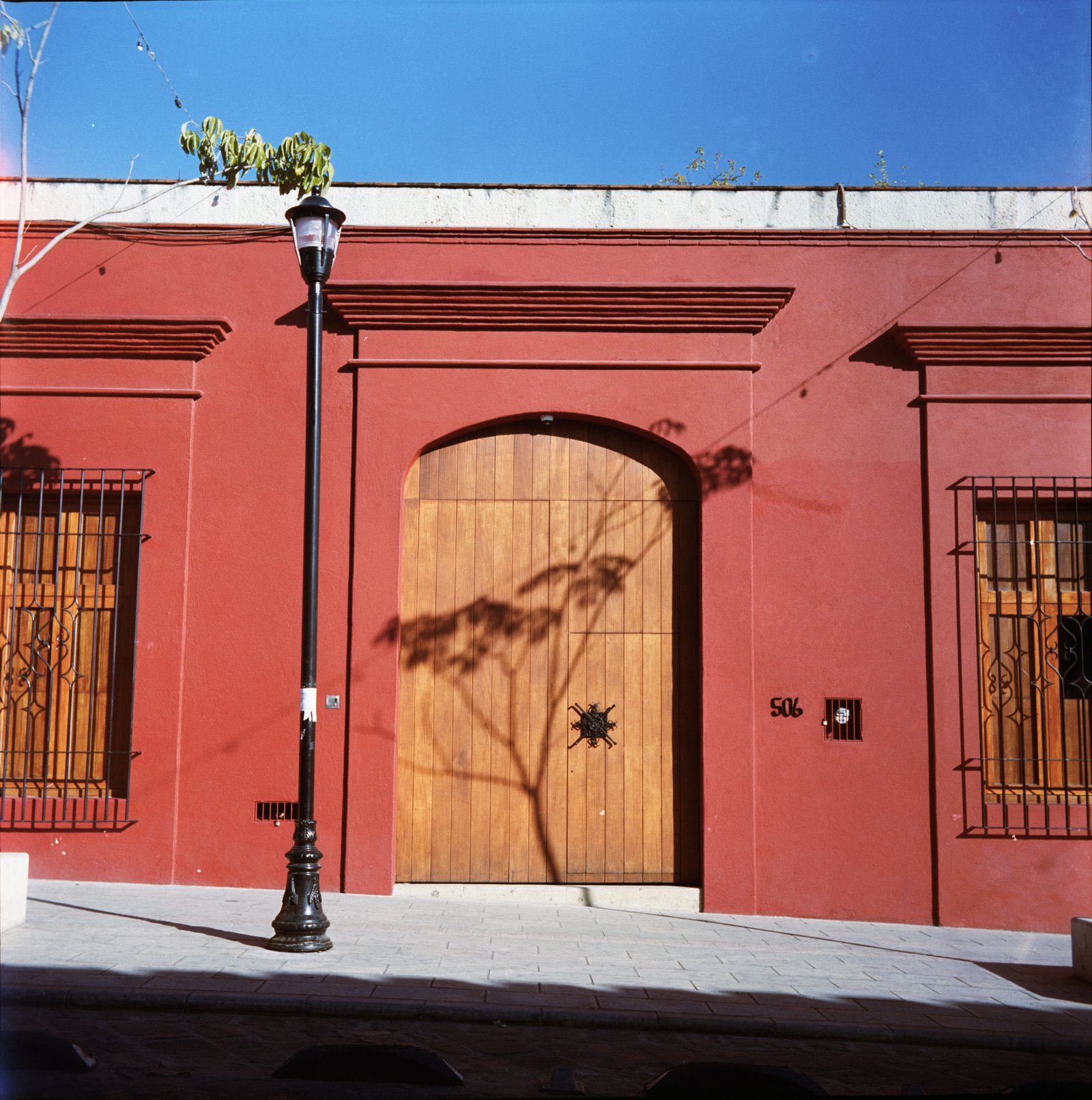 Copia de intiLab: fachada roja con una puerta de madera en arco, un farol y la sombra de un árbol.